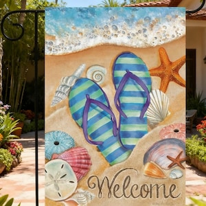 May include: A rectangular garden flag featuring a beach scene with blue and green striped flip-flops, various seashells, a starfish, and a sand dollar on tan sand. The word Welcome is written in a dark brown cursive script at the bottom.