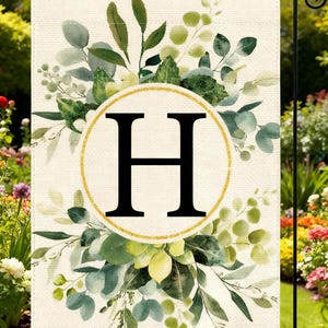 May include: A rectangular garden flag featuring a cream-colored background with a central gold-outlined circle containing a large black capital letter H. The design is surrounded by a watercolor-style arrangement of green eucalyptus leaves.