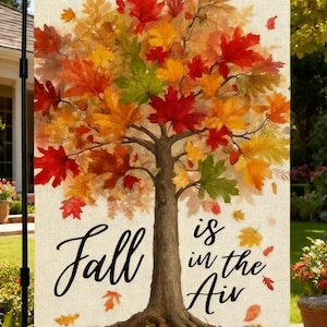 May include: A rectangular garden flag featuring a central tree with vibrant red, orange, yellow, and green autumn leaves. The text "Fall is in the Air" is printed in black script on the lower portion of the off-white, textured fabric background.