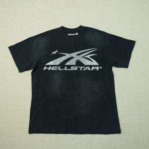 May include: A black short-sleeve t-shirt featuring a distressed silver graphic on the chest. The graphic displays a stylized star and swoosh design above the text HELLSTAR. A small brand label is visible on the inside of the crew neckline.