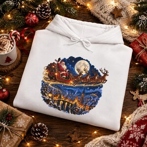 Può includere: Una felpa con cappuccio bianca caratterizzata da una stampa grafica circolare di Babbo Natale su una slitta trainata da renne che vola sopra un villaggio innevato di notte con una luna piena sullo sfondo. Cappuccio con coulisse.