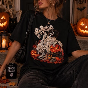 Può includere: Una t-shirt nera con la grafica di un fantasma che indossa un berretto e occhiali da sole, fuma ed è seduto tra zucche, foglie autunnali, un teschio e un gatto nero. Il fantasma è circondato da fumo e la maglietta è abbinata a jeans scuri.