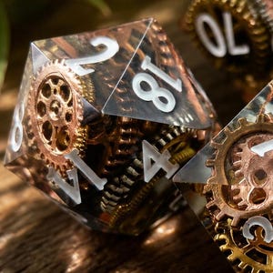 May include: A set of three polyhedral dice made of clear resin, each containing embedded metallic gear components in copper, brass, and silver tones. The dice feature white engraved numbers, including visible digits like 18, 40, 70, 50, 6, 5, 11, and 1.