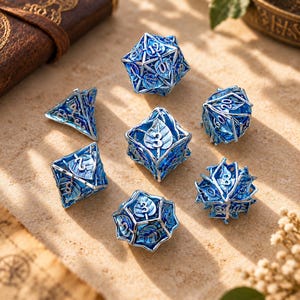 Op de afbeelding: Een set van zeven holle metalen veelzijdige dobbelstenen met een gedetailleerd zilverkleurig blad- en rankontwerp over een levendig blauwe emaille binnenkant. Elke dobbelsteen toont duidelijke, verhoogde cijfers, waaronder een twintig- en twaalfzijdige dobbelsteen.