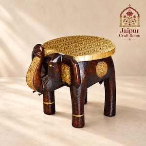 May include: A dark brown wooden stool shaped like an elephant with gold-colored accents on the ears, trunk, and legs. The flat, oval-shaped seat features a textured gold-colored top. The text Jaipur Craft Room is visible in the upper right corner.