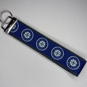 May include: A fabric keychain featuring a blue ribbon with a repeating Seattle Mariners compass logo pattern, stitched onto a dark gray webbing strap. The keychain includes a silver-toned metal hardware clamp and a circular split ring for keys.