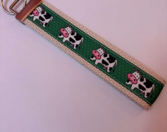 May include: A beige and green keychain wristlet featuring a repeating pattern of cartoon cows. The cows are black and white with pink accents. The keychain has a metal ring and a brown leather tab.