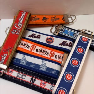 May include: A collection of fabric keychains featuring various Major League Baseball team logos and names. The keychains display team names like "Cardinals," "Mets," "Giants," "Dodgers," "Yankees," "Orioles," and "Cubs" in team colors.