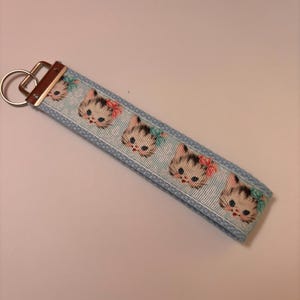 May include: A light blue key fob featuring a repeating pattern of vintage-style kitten faces with bows. The key fob has a metal key ring and a leather-like tab.