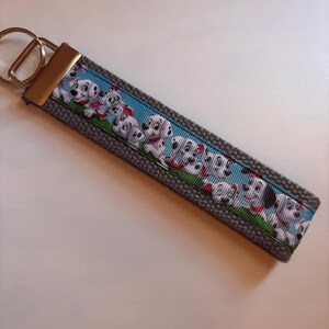 May include: A gray key chain with a blue and white ribbon featuring a pattern of Dalmatian puppies. The ribbon is sewn onto the gray fabric key chain.