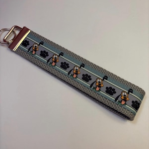 May include: Gray keychain wristlet featuring a repeating pattern of Pluto cartoon characters and black paw prints. The wristlet has a light blue stripe and a silver metal key ring.
