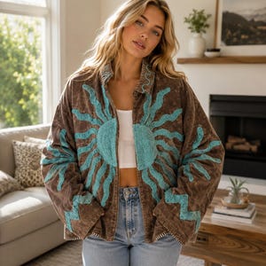 May include: A brown corduroy jacket featuring a large, embroidered turquoise sun design that spans across the front panels. The jacket has a collared neckline, long sleeves with zigzag embroidery, and a button-down front closure.