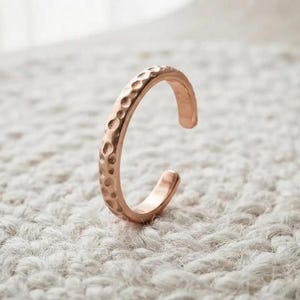 May include: A rose gold-toned open ring featuring a textured, hammered surface with small, circular indentations along the outer band. The ring has a C-shaped design with a gap, showcasing a polished metallic finish and a rounded, smooth inner edge.