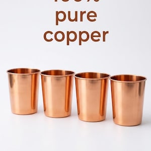 Puede incluir: Cuatro vasos cilíndricos de cobre pulido dispuestos en una fila horizontal. El texto 100% pure copper está impreso en marrón sobre los vasos. Cada vaso presenta un acabado metálico liso con un tono naranja-marrón cálido y uniforme.