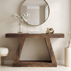 May include: A dark brown wooden console table with a trapezoidal base, featuring a clear glass vase with white orchid flowers, a stack of white books, and a gold-toned metallic knot sculpture on top, positioned below a round gold-framed mirror.