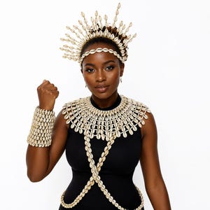 May include: A cowrie shell jewellery set consisting of a tall, radiating crown headpiece, a wide, multi-layered bib necklace that extends into a body chain, and a thick, cuff-style bracelet, all crafted from numerous small, off-white cowrie shells.