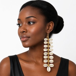 May include: A long, dangling earring featuring a vertical row of nine white cowrie shells arranged in pairs, connected by thin gold-toned wire. The earring has a symmetrical, linear design with a single shell at the bottom and a gold hook top.