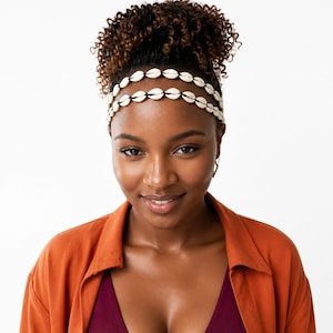May include: A double-strand headband featuring small, off-white cowrie shells strung along a thin black cord. The accessory is worn across the forehead, showcasing a repeating pattern of natural, oval-shaped shells against the dark cord material.