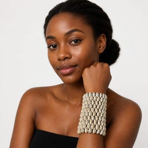 Puede incluir: Una pulsera ancha tipo brazalete compuesta por varias filas de pequeñas conchas de cauri de color blanco roto dispuestas en un patrón vertical. Las conchas están unidas firmemente, creando una banda cilíndrica texturizada alrededor de la muñeca.