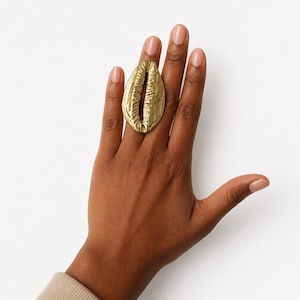 Puede incluir: Un anillo de tono dorado que presenta un diseño de concha de cauri grande y texturizada. La concha tiene una prominente ranura vertical central y un acabado de superficie rugoso y orgánico. El anillo se sitúa en el dedo medio.