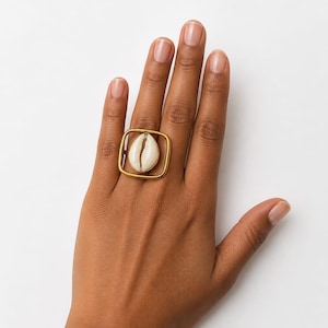 May include: A ring featuring a central white cowrie shell mounted within a square-shaped, matte gold-toned metal frame. The design showcases a smooth, rounded square band that encircles the shell, creating a geometric contrast with the organic form.