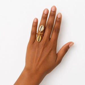 Puede incluir: Un anillo de tono dorado con dos elementos de concha cauri. El elemento superior es una concha cauri blanca natural, mientras que el elemento inferior es una réplica metálica de tono dorado. La banda es delgada y rodea el dedo en un bucle.