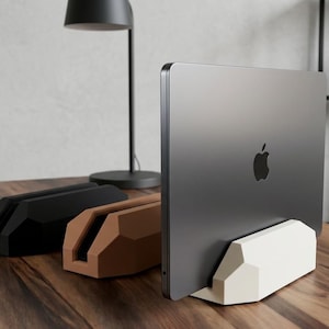 Op de afbeelding: Drie geometrische laptopstandaards in zwart, bruin en wit, elk met een centrale verticale gleuf. Een donkergrijze laptop met Apple-logo is in de witte standaard geplaatst, die op een houten oppervlak met zichtbare houtnerf staat.