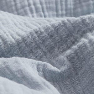 Op de afbeelding: Een lichtblauwe stof met een zichtbare, gekreukelde en meerlaagse textuur. Het materiaal heeft een rasterachtig weefpatroon met zachte plooien en schaduwen die diepte creëren over het oppervlak van het textiel, wat de lichtgewicht kwaliteit benadrukt.