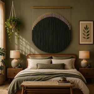 May include: A large circular wall hanging made of dark green fiber strands suspended from a horizontal wooden rod, featuring a row of light-colored wooden beads along the top edge, displayed above a bed with olive green and cream-colored bedding.