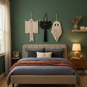 May include: Three macrame wall hangings featuring two off-white ghost figures and one black bat figure centered above a bed with gray bedding, a rust-colored throw blanket, and a cream-colored lumbar pillow with a spiderweb graphic.
