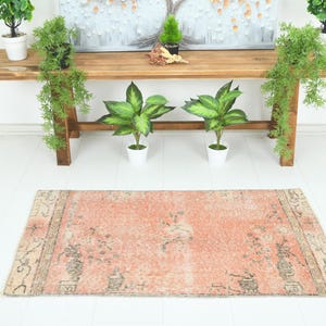 May include: A rectangular area rug featuring a faded coral orange base color with intricate, distressed floral and geometric patterns in muted gray and beige tones. The rug has a worn, vintage-inspired texture with a low pile and fringed edges.