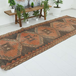 May include: A long rectangular runner rug featuring a repeating geometric pattern in shades of burnt orange and dark charcoal gray. The design consists of large central diamond motifs with intricate border details and angular lines throughout.