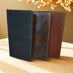 Puede incluir: Tres fundas rectangulares de cuero dispuestas verticalmente, con acabados en negro mate, marrón oscuro y marrón rojizo. Cada funda presenta costuras visibles de color claro a lo largo de los bordes, creando un aspecto limpio y estructurado.