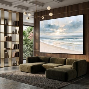 May include: A large rectangular wall art print featuring a serene coastal landscape with a sandy beach, gentle ocean waves, and a soft, cloudy sky in muted tones of blue, beige, and white, displayed in a black frame above a dark olive green sofa.