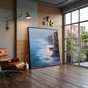 May include: A large framed landscape painting depicting a coastal scene with steep cliffs meeting the blue ocean, placed on a wooden floor next to a brown leather lounge chair, a small vase with dried branches, and a wall-mounted brass lamp.
