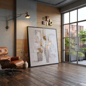 May include: A large square abstract painting with white, gray, and gold rectangular shapes in a black frame, positioned on a dark wood floor next to a brown leather lounge chair, a wall-mounted lamp, and a small vase with dried branches.