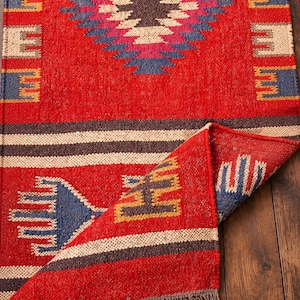 May include: A rectangular woven rug featuring a vibrant red base with a central diamond pattern in blue, pink, and cream. The rug includes horizontal stripes, geometric border motifs, and a folded corner revealing a brown fringe at the edge.