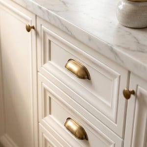 Può includere: Un mobile da cucina bianco con ante e cassetti a pannello incassato, dotato di maniglie a conchiglia e pomelli rotondi in ottone spazzolato. Un piano di lavoro in marmo bianco con venature grigie ospita un piccolo barattolo in ceramica maculata.