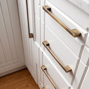 Puede incluir: Un juego de tiradores de armario de latón cepillado instalados en armarios de cocina blancos con paneles. Los herrajes presentan un diseño de barra rectangular con soportes cuadrados, proporcionando un contraste metálico sobre la madera blanca.