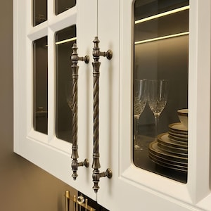 May include: White kitchen cabinet doors with glass panels and long, antique brass-toned metal handles featuring a twisted rope design. The interior of the cabinet displays stacked plates and crystal glasses, while the exterior features a black marble countertop.