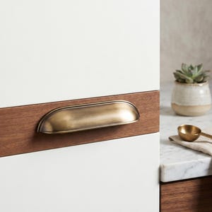 May include: A semi-circular cup drawer pull with a brushed antique brass finish, mounted horizontally on a flat wooden strip that is integrated into a white cabinet door. The metal handle features a smooth, curved design with a metallic sheen.