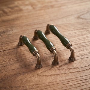 May include: Three cabinet pulls featuring a smooth, dark green ceramic center section with a glossy finish, connected to two curved, antique brass-toned metal mounting posts with a rounded base at each end of the handle.