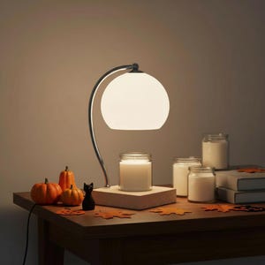 May include: A candle warmer lamp with a curved silver metal arm and a white glass dome shade. The lamp sits on a light wood base, illuminating a glass jar candle. Nearby are two small orange pumpkins, a black cat figurine, and two books.