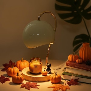 Puede incluir: Una lámpara calentadora de velas dorada con pantalla de cúpula blanca, colocada sobre una vela en frasco de vidrio sobre una base circular de madera. Incluye pequeñas calabazas naranjas, una figura de gato negro y hojas de otoño.