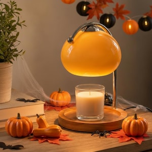 May include: A candle warmer lamp with a smooth, rounded orange glass shade and a silver metal arm, positioned over a clear glass jar containing a white wax candle on a circular wooden base, surrounded by small orange pumpkins and faux leaves.