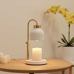 May include: A candle warmer lamp featuring a gold-toned metal arm and a matte white dome shade. The lamp sits on a circular white base, holding a white pillar candle directly beneath the light source, with a white power cord extending outward.