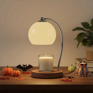 May include: A candle warmer lamp with a curved chrome arm and a spherical white glass shade. The lamp sits on a round wooden base holding a clear glass jar candle with white wax. The design features a minimalist aesthetic with a warm glow.