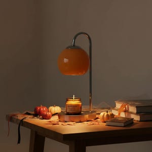 Puede incluir: Una lámpara calentadora de velas de metal con un brazo curvo y una pantalla esférica de color naranja brillante. La lámpara se asienta sobre una base que sostiene una vela en frasco de vidrio con una mecha encendida y calabazas decorativas.