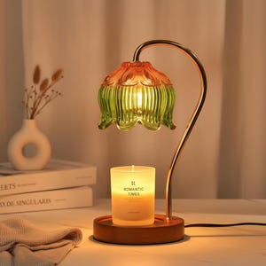 Może przedstawiać: Lampa do podgrzewania świec z zakrzywionym metalowym ramieniem w kolorze złota i szklanym kloszem w kształcie kwiatu z pomarańczowo-zielonym gradientem. Okrągła drewniana podstawa mieści białą świecę z napisem ROMANTIC TIMES na froncie.