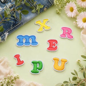 May include: A collection of seven individual embroidered letter patches in various colors including yellow, blue, red, pink, green, and orange. Each patch features a thick white border and a rounded, bold font style, arranged on a light green surface.